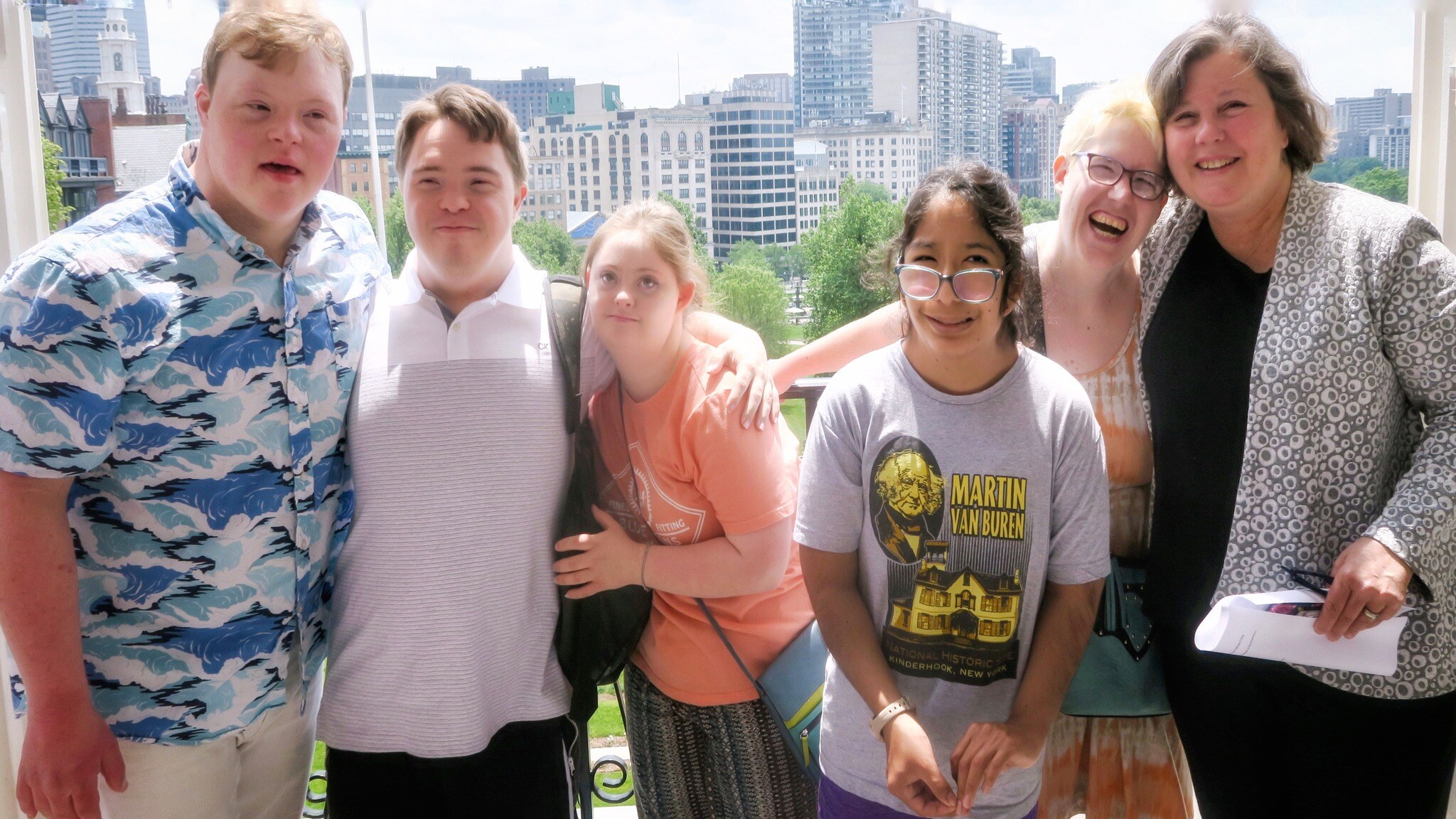A diverse group stands together, arms on each other’s shoulders, as they pose for a photo on the balcony of the Massachusetts State House.