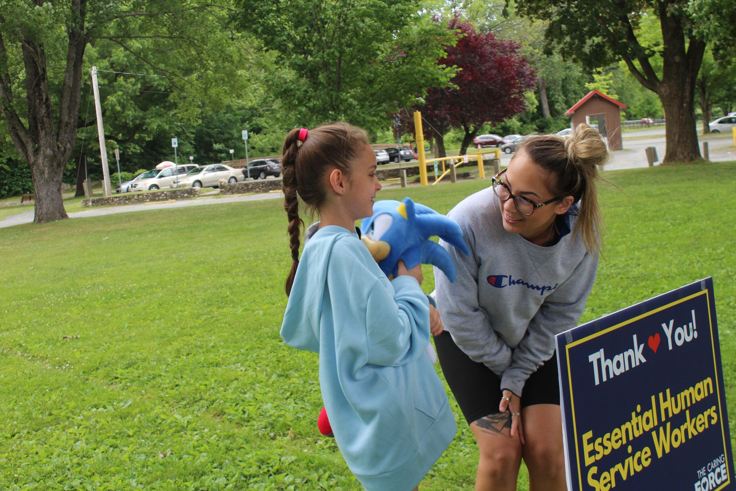 A young girl with a light skin tone and brown hair in a braided pony tail holds a sonic plush toy as she speaks with a young woman with tanned skin and blond hair in a ponytail. The woman is bending at her knees to speak with the girl.