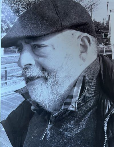 Black-and-white profile portrait of Michael Cooley, an older man with a disability, light skin tone, and short gray hair. He is wearing a flat-cap hat.