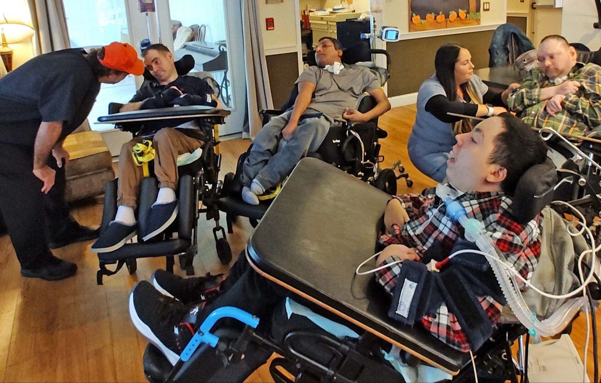 Four men with disabilities and diverse skin tones and ages sit in motorized wheelchairs. Some have tracheotomy tubes attached to the back of their wheelchairs. Two nurses each speak to one of the men in the wheelchairs.