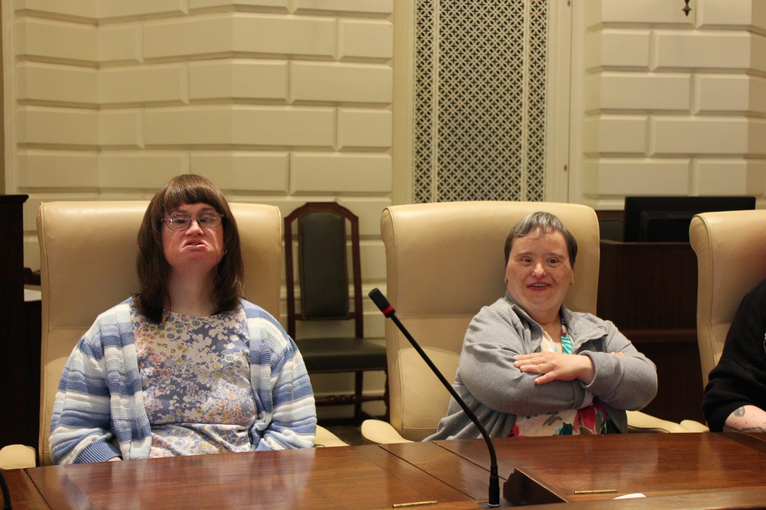 Two women with disabilities and light skin tones sit in the chairs of senators at the Massachusetts State House.