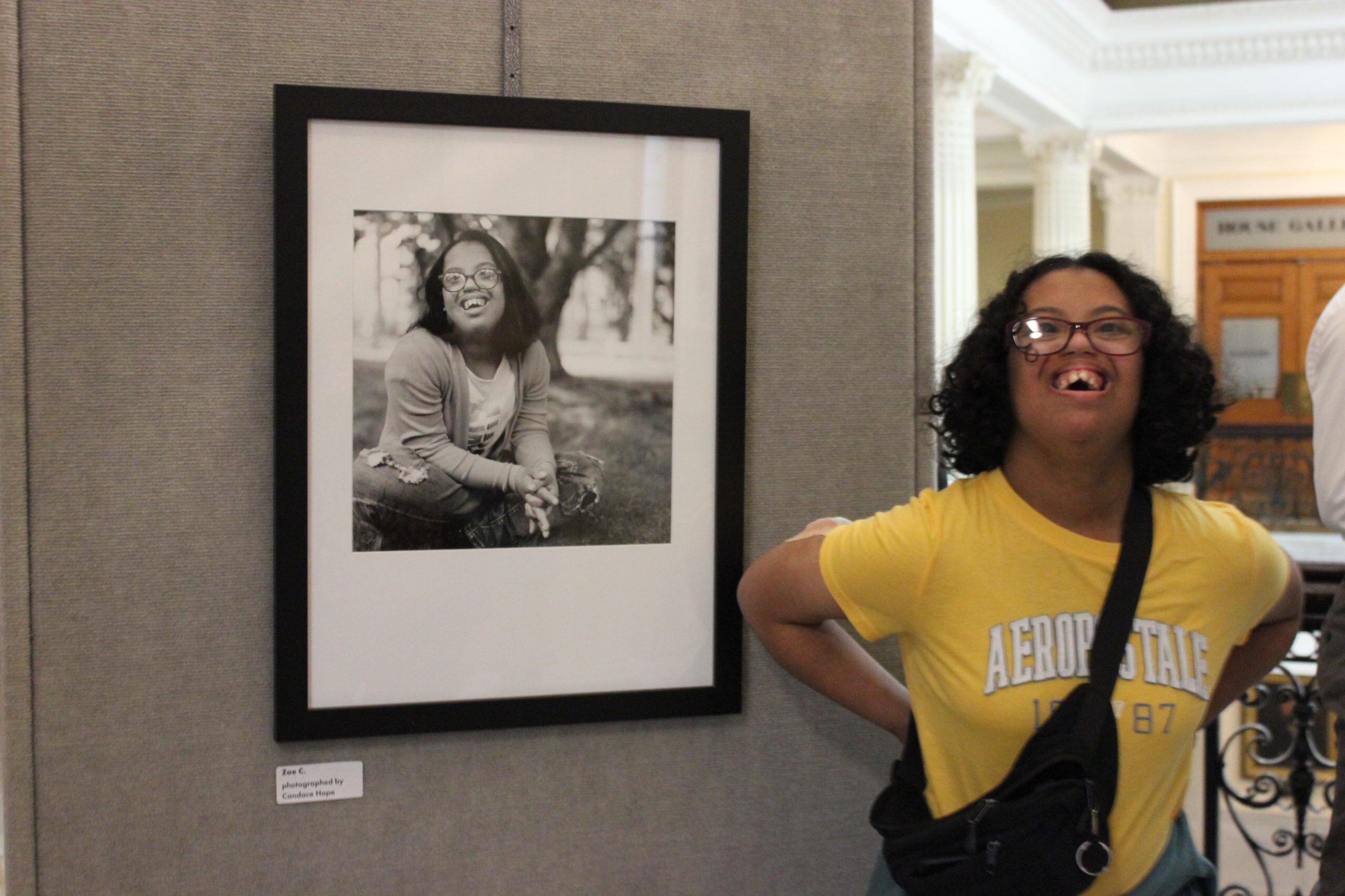 Zoe Cummings, a woman with a disability, a dark skin tone, and chin-length curly black hair, laughs in front of a black-and-white photo of herself.