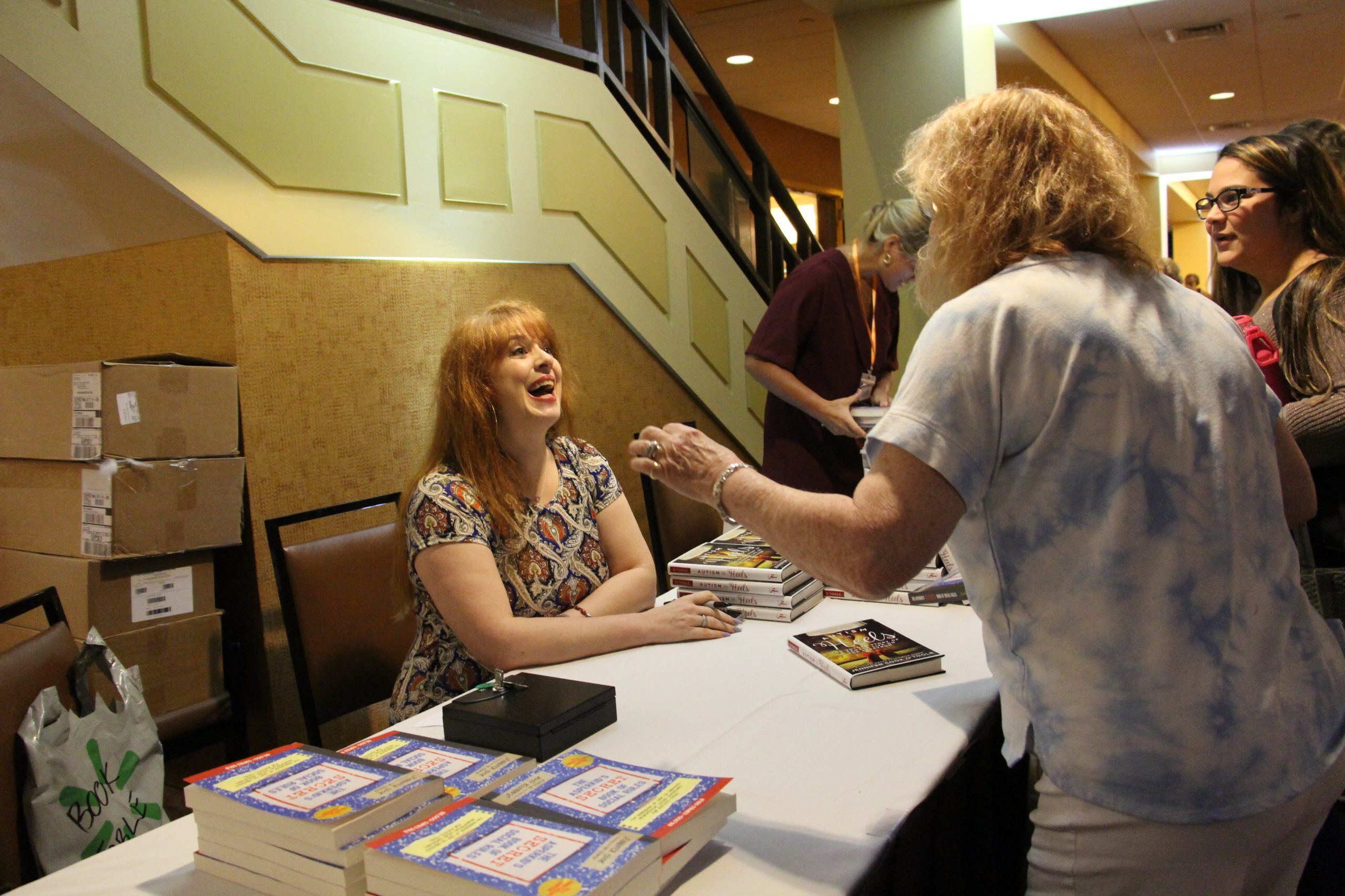 Jennifer Cook, a woman with a light skin tone and long red hair, sits at a booth while happily speaking with an older man with a light skin tone and chin-length blond hair.