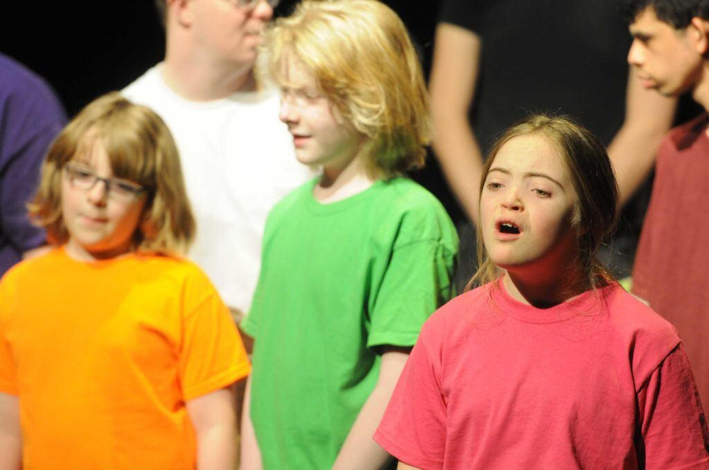 Whole Children and Milestone. A young girl with a disability and light skin tone is wearing a red shirt while singing West Side of the Bridge in the foreground. Behind her are other children wearing different-colored shirts.